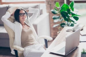 A woman leans back at her computer desk in a comfortable office chair. She is wearing white.