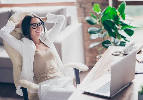 A woman leans back at her computer desk in a comfortable office chair. She is wearing white.